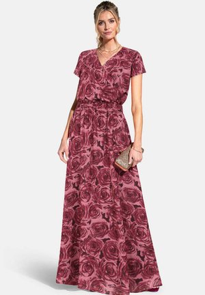 Žena v dlhých šatách s potlačou ruží a krátkymi rukávmi, drží malú trblietavú kabelku v pravej ruke.