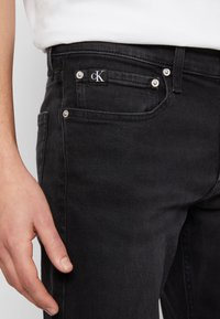 Black denim jeans featuring a cK label, silver rivets, and a classic five-pocket design. Material has a smooth, slightly textured finish.