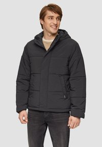 Schwarze Daunenjacke mit Kapuze, mit gestepptem Design, Vordertaschen und Reißverschluss. Getragen über einem beigen Pullover und dunklen Jeans.