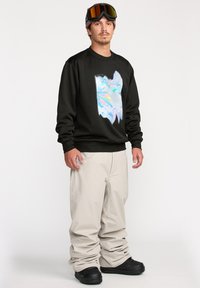 Sweatshirt noir avec un design abstrait coloré sur le devant, associé à un pantalon beige ample et des bottes de neige noires.