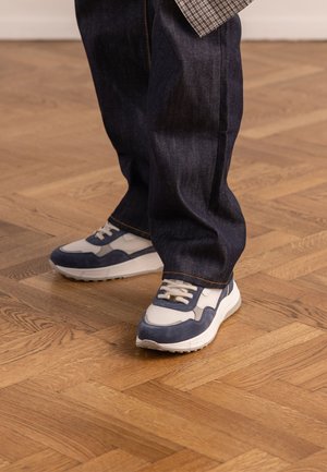 Persoon met witte en marineblauwe sneakers met witte veters en donkerblauwe spijkerbroek, staand op een houten parketvloer.