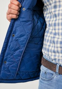 Inderside af blå quiltet jakke med trykknapper, der viser en glat tekstur. Bærtes over en ternet skjorte og blå denim med et brunt bælte.