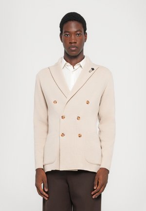 Beige double-breasted blazer made of textured fabric featuring large brown buttons, a shawl collar, and patch pockets. Black accent pin included.