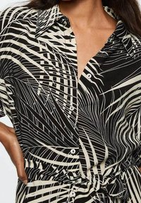 Zwart-wit shirt met een gestreept patroon dat lijkt op bladeren, korte mouwen, een kraag en knoopsluitingen; getailleerde taille met strikdetail.