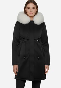 Parka nera con colletto in pelliccia bianca, dotata di zip frontale, cordini regolabili e due tasche laterali. Tessuto liscio e resistente all'acqua.