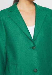 Blazer verde de mezcla de lana con cuello de muesca, mangas largas, dos bolsillos delanteros y dos botones negros en la parte frontal. Textura suave.