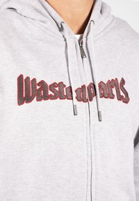 Grå zip-opp hettegenser laget av mykt stoff, med rød og svart brodert tekst "Wastedparts" på brystet. Inkluderer justerbare snører.