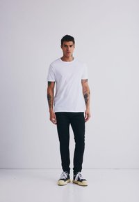 Valge lühikeste varrukatega t-särk, mis on kokku sobitatud mustade slim-fit pükstega ja musta-valgete spordijalatsitega, taustal ühtlane taust.