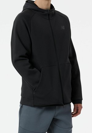 Personne portant un sweat à capuche noir zippé avec des poches et un petit logo sur la poitrine, associé à un pantalon bleu-gris.