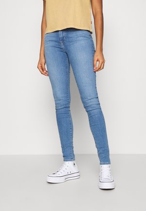 Vaqueros ajustados en denim azul claro; presentan una textura suave, costuras mínimas y una cintura de altura media. Combinados con zapatillas blancas.