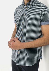 Camicia a maniche corte con bottoni in motivo gingham bianco e nero, con dettagli in denim blu su colletto e maniche. Presenta una tasca sul petto.