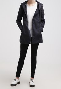 Chaqueta con capucha de color navy, con cierre de cremallera, bolsillos delanteros y tejido ligero, llevada sobre una camiseta blanca y combinada con pantalones negros y zapatillas blancas.