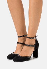 Gambe femminili che indossano scarpe con tacchi alti in suede nero con doppie cinghie alla caviglia e fibbia argentata su uno sfondo bianco.
