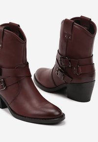 Des bottes en cuir bordeaux montantes présentent un bout pointu, un talon empilé et des accents de double boucle sur le côté pour un design structuré.