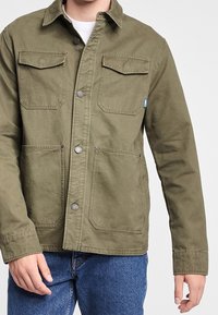 Olive green canvas jacket featuring a front button closure, two chest pockets, and two side pockets, with a relaxed fit and minimal stitching.