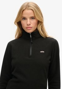 Zwarte fleece pullover met een hoge kraag, halve ritssluiting en kleine branding op de linkerborst. Zacht, gestructureerd materiaal.
