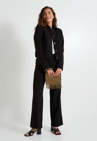 Mujer con chaqueta y pantalones negros, sosteniendo un bolso de flecos dorados, sonriendo y mirando de lado contra un fondo blanco.