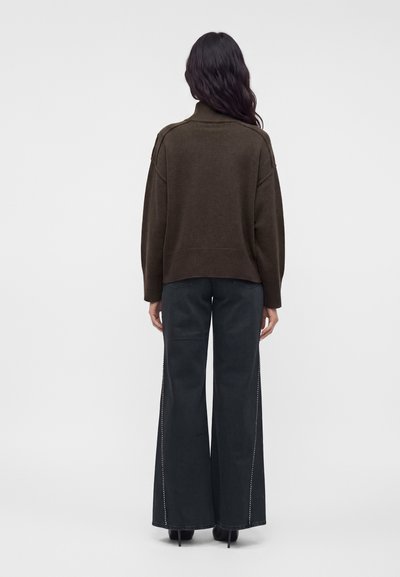 Femme aux longs cheveux bruns foncés portant un pull marron foncé et un pantalon noir à larges jambes, debout, dos à un fond uni.