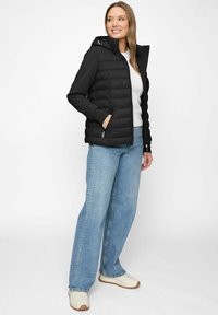 Woman wearing black hooded quilted jacket, white shirt, wide-leg blue jeans, and white sneakers, standing and smiling, white background.