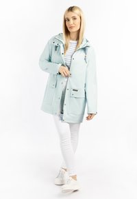 Világoskék vízálló dzseki kapucnival, ezüstszínű fémrészekkel, elülső zsebekkel és cipzárral. Csíkos póló fölé van húzva, fehér nadrággal párosítva.