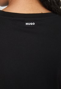 Gros plan du col arrière d'une chemise noire avec le texte blanc "HUGO" imprimé sous le col sur une personne aux cheveux foncés.