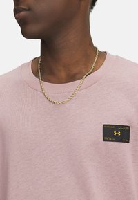 Light pink cotton T-shirt featuring a black label with yellow text on the left chest and a crew neckline. Gold twisted chain necklace visible.