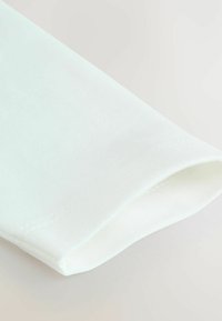 Tela de tejido blanco con una textura suave y lisa, que presenta un borde enrollado y un detalle sutil de costura a lo largo del dobladillo.