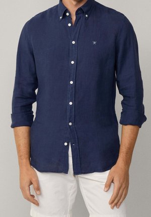 Homme portant une chemise bleu marine à manches retroussées et un pantalon blanc, debout les bras détendus le long du corps.