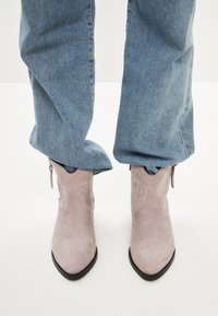 Bocanci cowboy din suede gri deschis, cu vârfuri ascuțite, cusături decorative și fermoare laterale, asortați cu blugi din denim albastru.