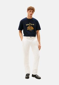 Joven de pie, vistiendo una camiseta gráfica azul marino, pantalones blancos y mocasines negros, contra un fondo claro y liso.