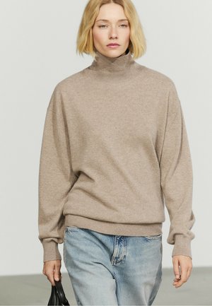 Lyshåret kvinde iført en beige rullekravesweater og lyseblå jeans, der holder en lille sort håndtaske i venstre hånd.