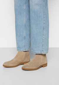Stivaletti Chelsea in suede beige con pannelli elastici laterali, tacco basso in legno e suola liscia color paglia, abbinati a jeans azzurri chiari arrotolati.