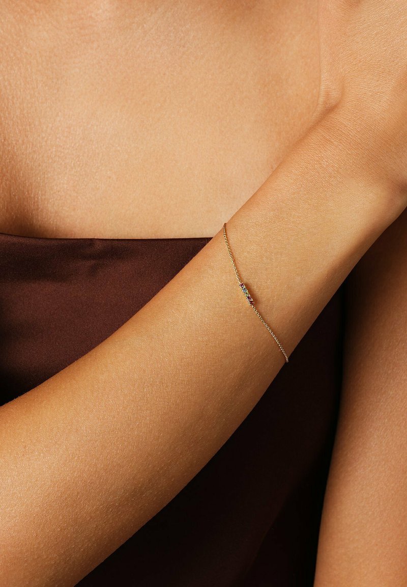 Sakral guldkedja med små flerfärgade ädelstenar som bärs på en upphöjd underarm mot en brun tygbakgrund.