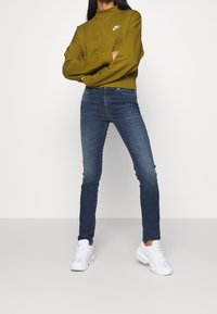 Osoba v olivově zelené mikině Nike, modrých skinny džínách a bílých teniskách stojící s překříženýma rukama na jednobarevném pozadí.