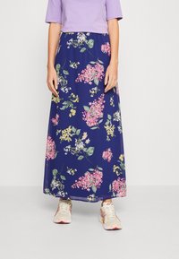 Kvinna i en lavendelblå topp, marinblå maxikjol med rosa och gula blommönster, och vita sneakers som står på ett ljust grått golv.