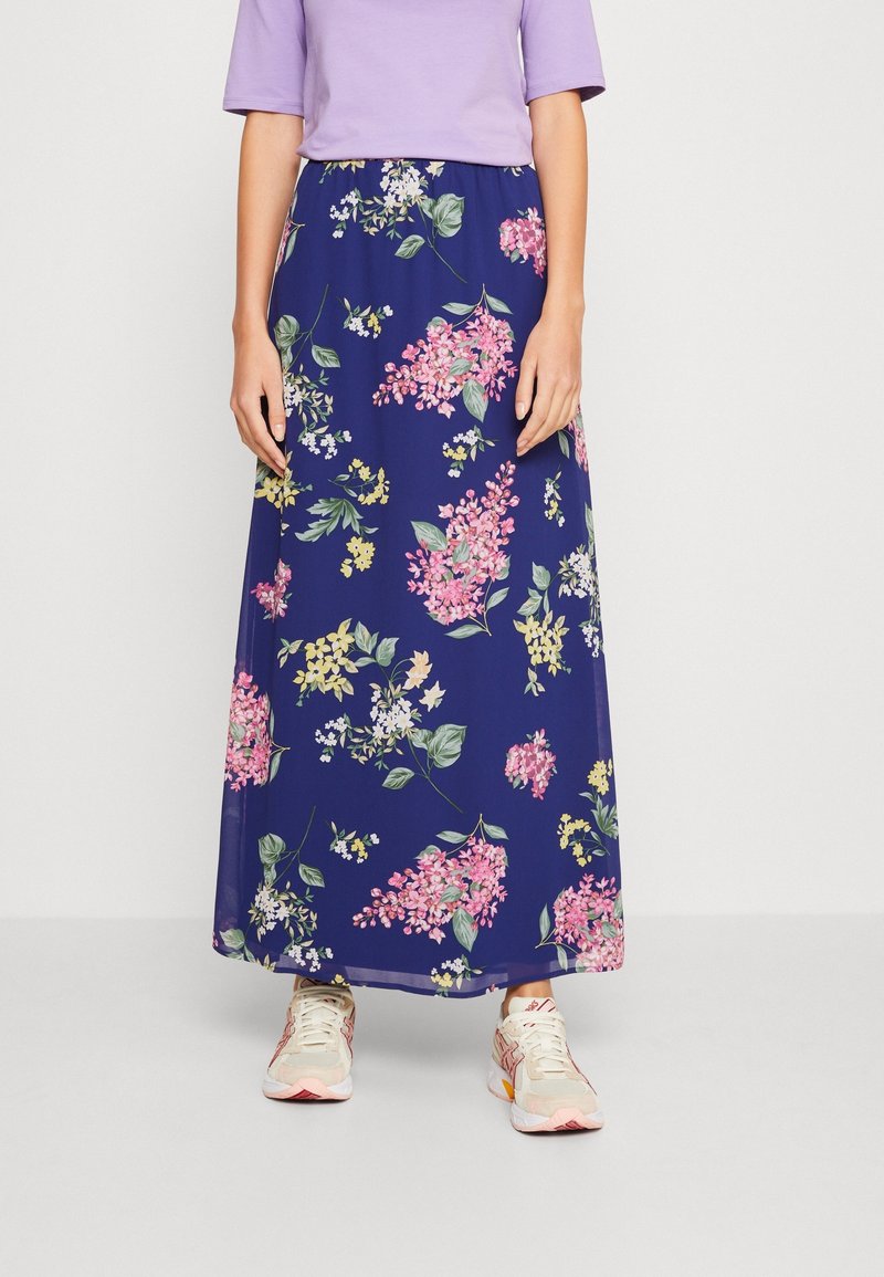 Kvinna i en lavendelblå topp, marinblå maxikjol med rosa och gula blommönster, och vita sneakers som står på ett ljust grått golv.