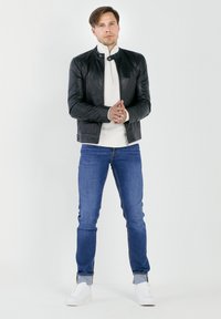 Zwarte leren jas met een opstaande kraag, witte gebreide trui, blauwe spijkerbroek en witte sneakers. Model staat met handen gekruist.