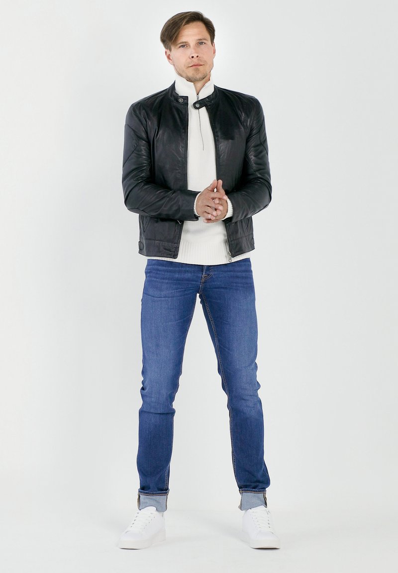 Zwarte leren jas met een opstaande kraag, witte gebreide trui, blauwe spijkerbroek en witte sneakers. Model staat met handen gekruist.
