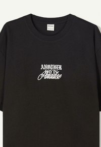 Svart bomulls t-shirt med en vit grafisk textdesign som säger "EN ANNAN DAG I PARADISET" på bröstet. Korta ärmar och rund hals.