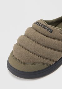 Maslinasto zeleni papuč s ušivenim dizajnom i crnim gumenim potplatom. Sadrži teksturirani materijal i logo "HILFIGER" u mornarsko plavoj boji na gornjem dijelu.