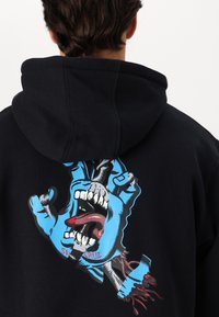 Melnā kapuces krekls ar lielu zilu zīmējumu, kas attēlo roku ar atvērtu muti, izrādot zobus un mēli, ar sarkaniem akcentiem ap dizainu.