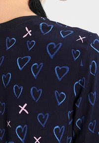Dunkles Gewebe mit einem Muster aus blauen Herzen und rosa Xs in verschiedenen Größen, mit einer weichen Textur und rundem Ausschnitt.