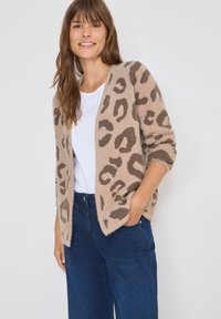 Luddig beige kofta med brunt leopardmönster, öppen front och trekvartslånga ärmar, bärs över en vit skjorta och blåjeans.