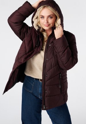 Blonde vrouw draagt een donkerbruine pufferjas met capuchon, beige trui en blauwe spijkerbroek, houdt de capuchon vast en kijkt naar de camera.