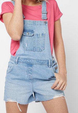 Personne portant une salopette courte en denim bleu clair et un t-shirt rose à manches courtes, une main dans la poche et l'autre près du visage.