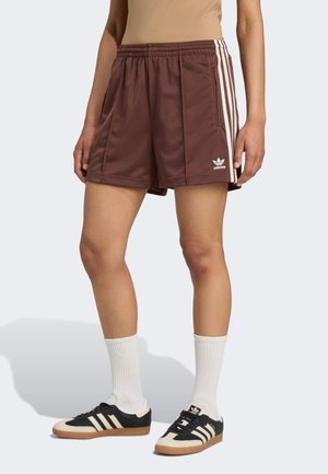 Persoon draagt bruine Adidas-shorts met witte strepen, crème en zwarte Adidas-sneakers en witte sokken, staand op een lichte achtergrond.