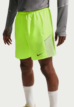 Mies, jolla on päällään neonkeltaiset urheilushortsit sivuvärttinäverkotaskulla ja harmaa paita, käsivarret ja jalat näkyvissä reiden puolivälistä käsiin, valkoiset sukat.