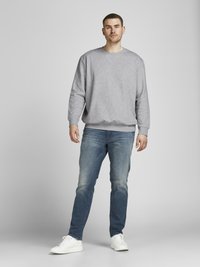 Felpa grigia con scollo rotondo, abbinata a jeans blu sbiaditi e sneaker bianche, in piedi contro uno sfondo neutro.