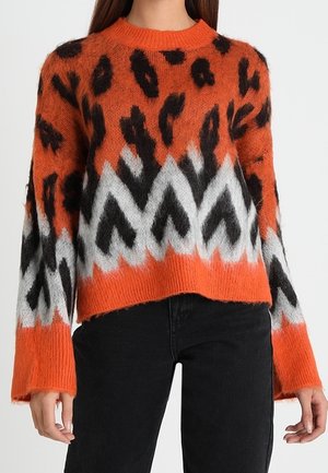 Pull orange pelucheux avec des taches léopard noires et un motif en zigzag blanc, porté avec un pantalon noir.