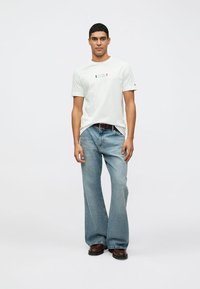 Balta kokvilnas T-kreklu ar centrā izdrukātu logo, apvienota ar gaiši zilām platiem džinsiem. Modelis valkā tumšas kurpes, stāvot pret neitrālu fona.